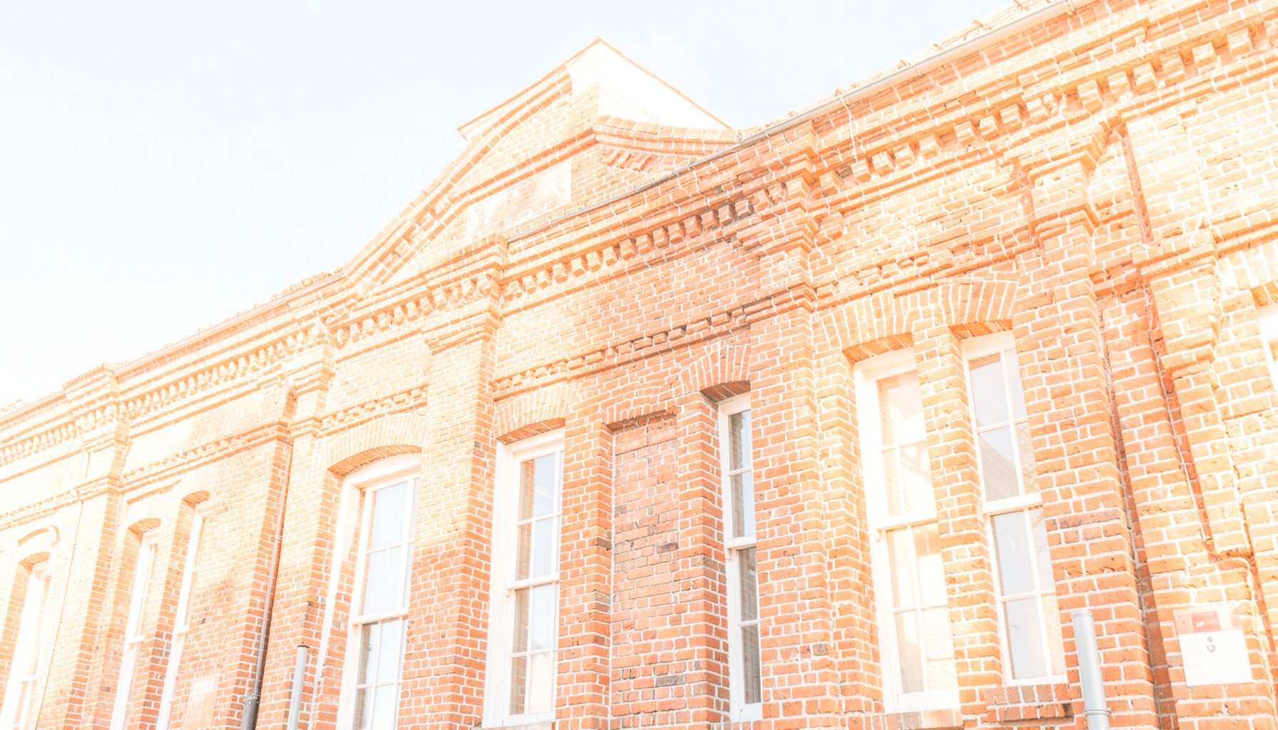 A bright, airy photograph of a historic brick building facade under a clear sky with warm sunlight casting gentle shadow