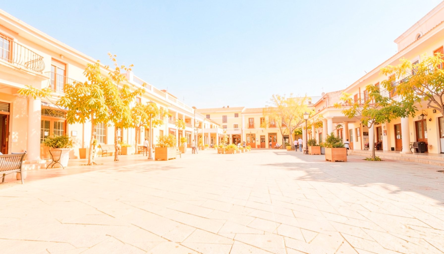 A bright, airy wide photograph of a sunlit open plaza with warm golden-hour light, soft shadows, and a clear blue sky
